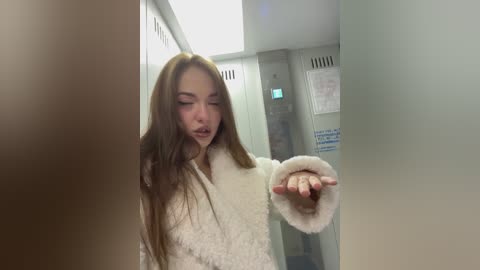 Media: Video of a young woman with long brown hair, wearing a white, fluffy bathrobe, standing in a dimly lit locker room. She is looking down and touching a white towel with her right hand.