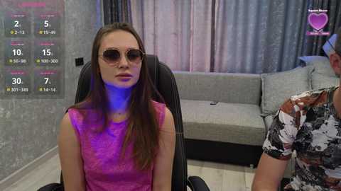 Video of a young woman with long brown hair, wearing a pink lace top and sunglasses, sitting in a black office chair. Background includes a gray couch, a textured gray wall, and a digital display showing statistics.