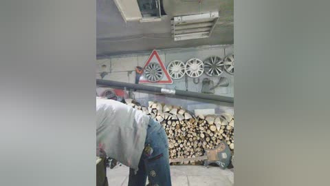 Media: Video of a man in a blue shirt, jeans, and a cap, bending over to grab a log, in a dimly lit, cluttered workshop with a pile of stacked logs, a red triangular road sign, and several ceiling vents.