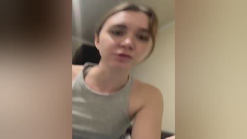 Media: A blurry video of a young woman with light skin, brown hair, and a gray tank top, taken from a low angle, showing a close-up of her face and upper body.