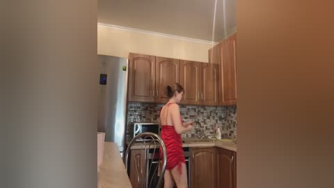 Media: Video of a woman in a red dress, standing in a modern kitchen with wooden cabinets and a mosaic backsplash, preparing food at the counter.