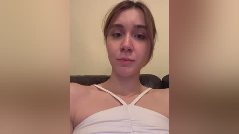 Media: A video of a young woman with light skin, brown hair tied back, wearing a white spaghetti-strap top, sitting on a brown couch against a beige wall.