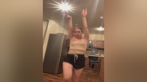 Media: Video of a woman in a beige tank top and black shorts, dancing in a dimly lit kitchen with wooden floors, a fridge, and cabinets.