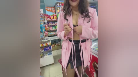 Media: Video of a dark-skinned woman in a pink robe with a black lace bra and high-waisted shorts, standing in a brightly lit convenience store with colorful candy displays and shelves.