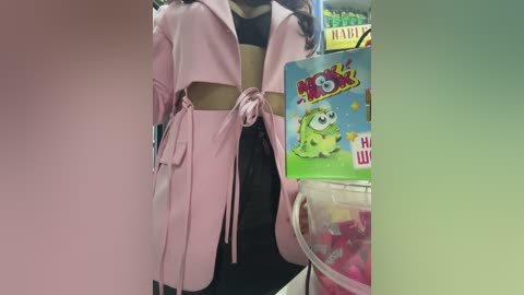 Media: Video of a person wearing a pink trench coat with a black belt and black pants, holding a green plush toy in a store setting.