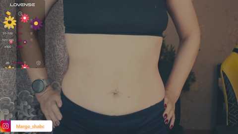 Video of a light-skinned woman wearing a black sports bra and black pants, with a visible navel and a tattoo on her right arm, standing against a patterned background.