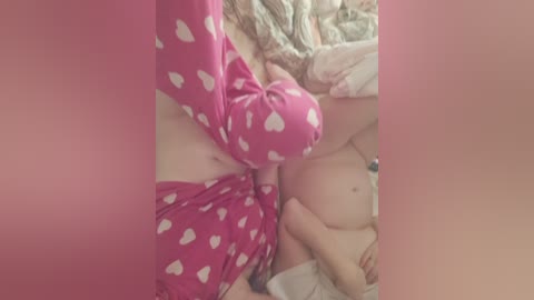 A close-up video of a woman lying on a bed, wearing pink heart-patterned pajamas, her hand touching her exposed breasts. The background shows a mix of pastel-colored sheets.