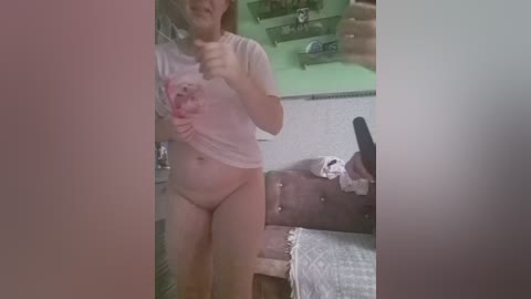A candid photo of a young woman in a white T-shirt and no pants, holding a phone in a dimly lit room with a green ceiling and a bed in the background.