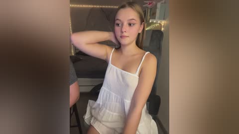 Media: Video of a young Caucasian girl with light skin, brown hair, and a slender physique, wearing a white, thin-strapped dress, sitting on a chair in a dimly lit room with string lights and dark furniture.