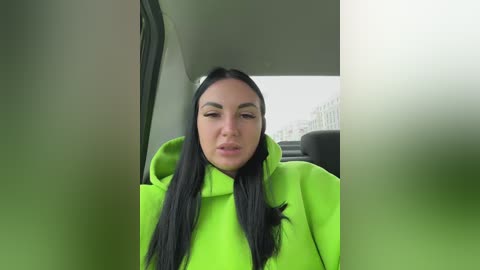 Video of a young woman with long black hair and light skin, wearing a bright green hoodie, seated in a car with a blurred cityscape visible through the window.