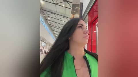 Video of a young woman with long black hair, wearing a bright green jacket, walking in a modern, bright mall with red and white accents.