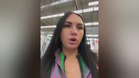 Video of a young woman with long black hair, wearing a colorful plaid shirt, standing in a dimly lit, industrial building with exposed ceiling beams and fluorescent lights.