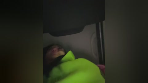 A dimly-lit video of a person lying on the ground, wearing a bright yellow jacket, partially covered by a black object, with a blurred background.