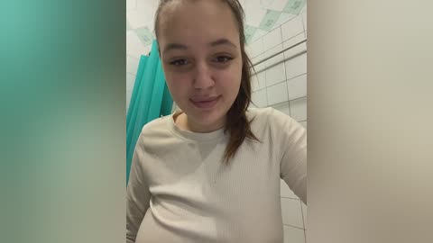 Media: Video of a young woman with fair skin, brown hair, and a slim build, wearing a white long-sleeve shirt, standing in a bathroom with white tiled walls and a turquoise shower curtain.