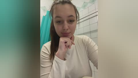 Media: A video of a young woman with fair skin and brown hair, wearing a long-sleeved white shirt, leaning on a bathroom wall, with a tiled shower in the background.