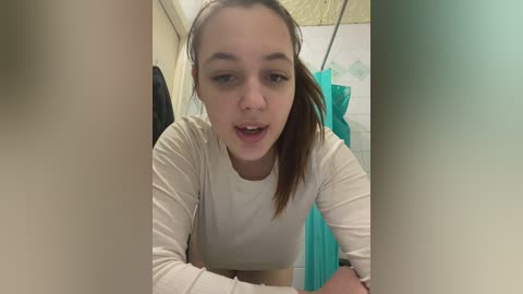 Media: Video of a young Caucasian woman with fair skin and brown hair tied back, wearing a white long-sleeve shirt. She is leaning over a sink in a bathroom with white tiles and a teal shower curtain.