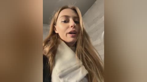 Media: Video of a Caucasian woman with long, blonde hair, wearing a white scarf, against a light brick wall, indoors, in a slightly blurred, candid selfie.