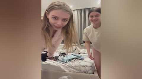 Media: A video captures two young women with light skin and long blonde hair, one in a pink sweater, the other in beige, standing beside a messy bed in a dimly lit bedroom.