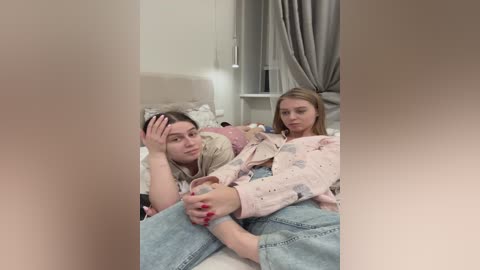 Media: Video of a young woman with light skin and straight blonde hair, wearing a pastel floral top, holding a teddy bear. She lies in bed with a young man, both looking concerned, in a dimly lit room with beige walls and curtains.