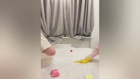 Media: A video of a person in a hospital room, wearing a white robe and yellow gloves, cleaning a white bed with pink and yellow cleaning cloths, gray curtains in the background.