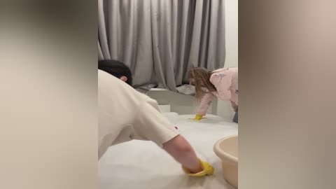 Media: Video of two women in white aprons and yellow gloves painting a white wall. One woman has long, blonde hair, while the other has short, dark hair. Gray curtains in the background.