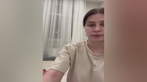 Media: Video of a young woman with fair skin and dark brown hair, wearing a beige T-shirt, standing beside a tall, beige curtain in a room with white walls and a window with sheer curtains.