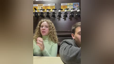 Media: Video of a young woman with curly hair, wearing a light green turtleneck, and a man with short brown hair, both sitting in a booth with a wall of toy dolls and boxes.