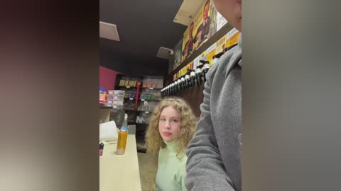 Media: Video of a young blonde woman in a green turtleneck, seated in a dimly lit bar, holding a beer, with a man in a gray shirt standing behind her.