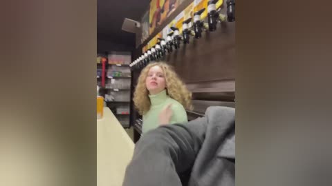 Media: Video of a woman with curly blonde hair, wearing a light green turtleneck sweater, seated at a booth in a dimly lit, cluttered room with shelves and a colorful mural on the wall.