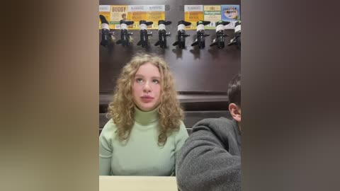 Media: Video of a young woman with curly blonde hair, wearing a light green turtleneck, seated in a dimly lit, high-backed booth with multiple microphones. Background features a menu board and other patrons.