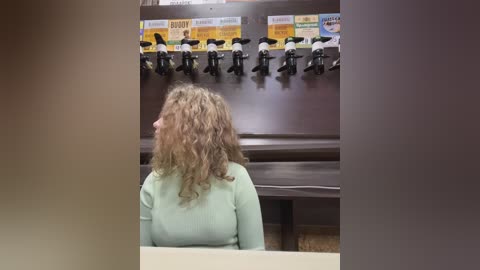 Media: Video of a person with curly brown hair, wearing a light green sweater, sitting at a bar with multiple beer taps and labeled kegs in the background.