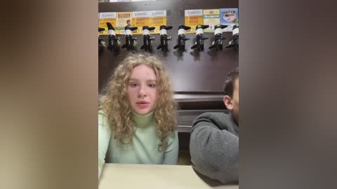 Media: Video of a curly-haired young woman in a mint green sweater, seated at a booth in a diner, with a man in a gray hoodie partially visible.