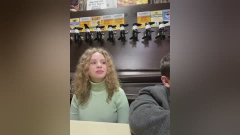 Media: Video of a young girl with curly blonde hair wearing a light green turtleneck sweater seated at a wooden table in a dimly lit room, with a man in a grey hoodie in the background.
