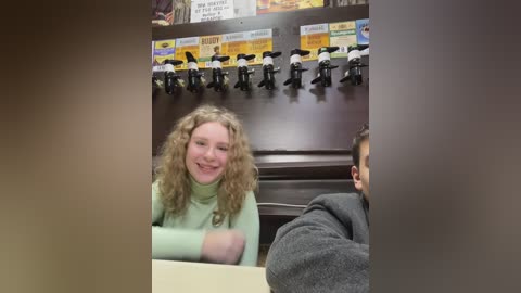 Media: Video of a young woman with curly hair, smiling, seated at a counter in a diner. Behind her, a row of black and white vintage microphone stands hangs from the ceiling.
