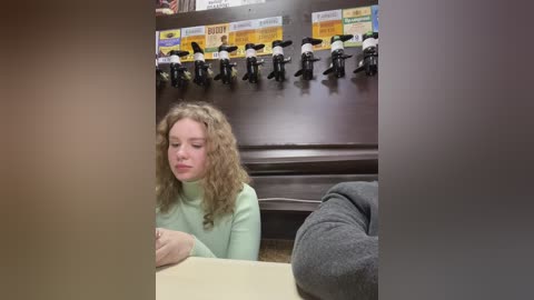Media: Video of a young girl with curly hair, wearing a light green sweater, sitting at a table in a cafeteria. Behind her, a shelf displays toy dolls and signs.
