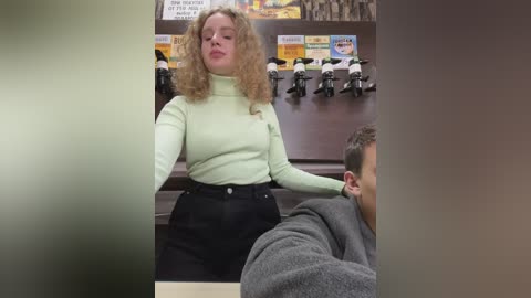 Media: Video of a blonde woman in a mint green turtleneck, black high-waisted pants, leaning against a counter in a barbershop with multiple black and white motorbike figurines in the background.