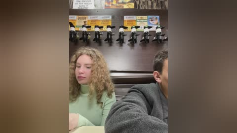 Media: A video of a young woman with curly hair in a green sweater, seated beside a man in a gray sweater, in a hospital waiting room with toy police cars and a \"Barnes & Noble\" poster on the wall.
