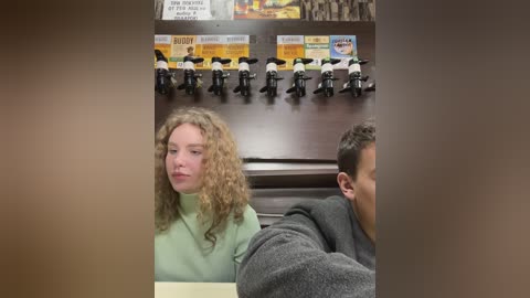 Media: Video of a curly-haired woman in a green sweater seated in a restaurant booth. Behind her, a table displays a LEGO police setup, including toy motorcycles and a sign.