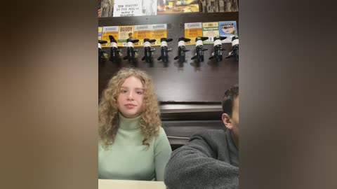 Media: Video of a young woman with curly blonde hair and a light green turtleneck sweater, seated in a restaurant with dark wooden walls and mounted microphones, reading a menu.