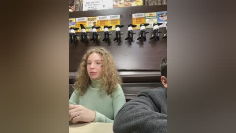 Media: Video of a young girl with curly hair in a green sweater, sitting at a booth in a fast-food restaurant. Behind her, a display case holds toy figures.