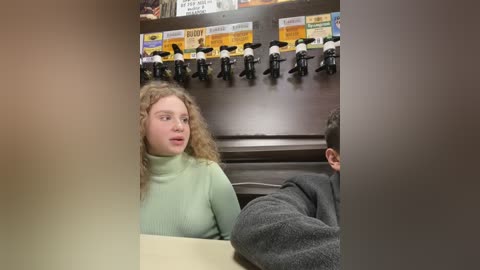 Media: A video of a young woman with curly hair in a mint-green turtleneck sweater, seated in a coffee shop. The background features a row of black coffee machines and a menu board.