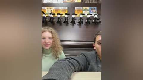 Media: Video of two people, a curly-haired woman in a green sweater and a man in a gray sweater, seated in a coffee shop with a wooden counter and a row of coffee dispensers in the background.