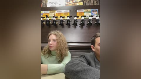 Media: Video of a blonde woman with curly hair in a green sweater and a man in a gray shirt, sitting at a table in a restaurant with a menu board and toy motorcycles on the wall.