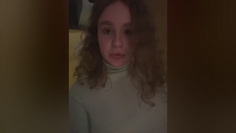 Media: A dimly lit video of a young woman with curly brown hair and pale skin, wearing a beige turtleneck sweater, standing against a dark, indistinct background.