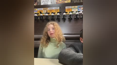 Media: Video of a woman with curly blonde hair in a green turtleneck sweater seated at a counter, surrounded by a wall of \"Burger King\" posters and toy horse figurines.