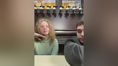 Media: Video of a curly-haired woman in a green sweater and a man in a grey hoodie sitting side-by-side at a table, with a wall-mounted row of bicycles behind them.