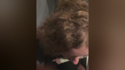 Media: Video of a man with curly, light brown hair, slightly bent forward, wearing a black shirt, and holding a light-colored object in his hand, blurred background.
