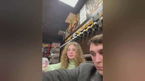 Media: Video of two young people, a blonde girl in a green sweater and a boy with short hair, sitting in a dimly lit arcade. The background features a colorful mural and multiple arcade machines.