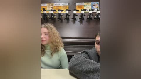 Media: Video of a woman with curly hair and a green turtleneck, seated at a table in a diner, surrounded by a wall display of black-and-white photos.
