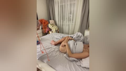 Media: Video of a woman lying on a messy bed, wearing a gray sweater, with a phone in hand, surrounded by a disheveled room with a window and floral decorations.